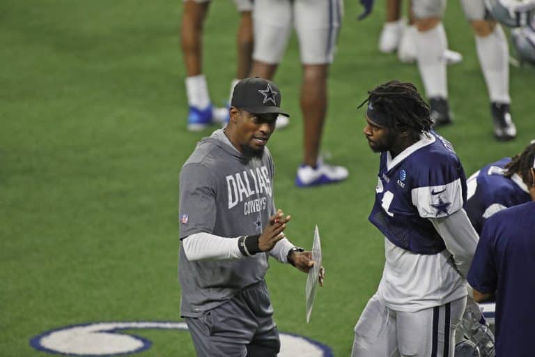 Dallas Cowboys coach in a grey outfit talks to player Trevon Diggs on the field with others in the background.