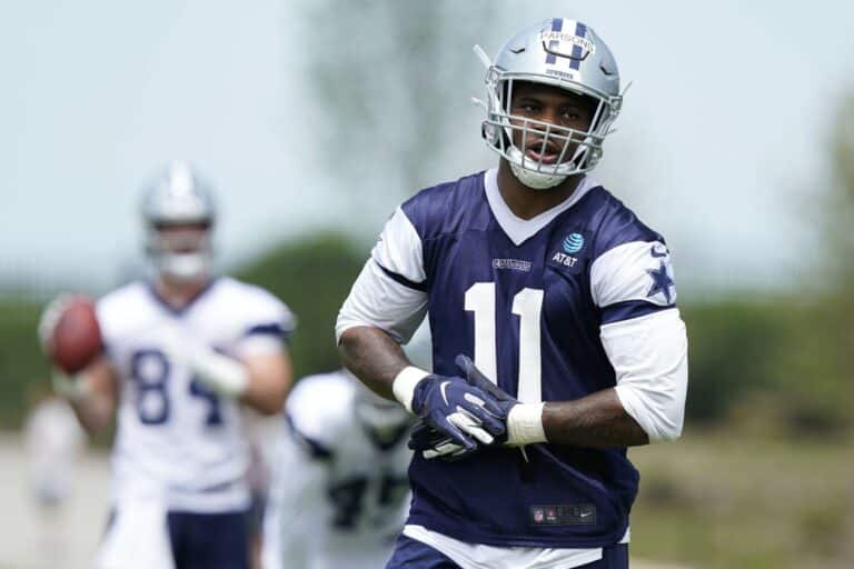 A football player in a navy and white uniform practices at Training Camp, with a teammate holding a football in the background.