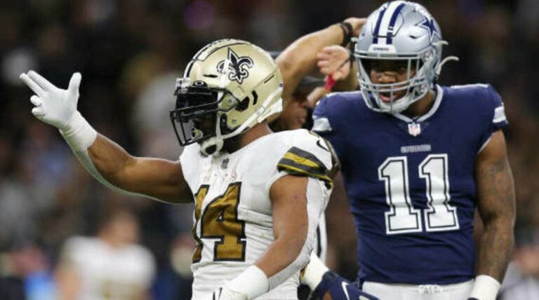 A Saints football player in a white and gold uniform celebrates while a player in a blue uniform stands in the background.