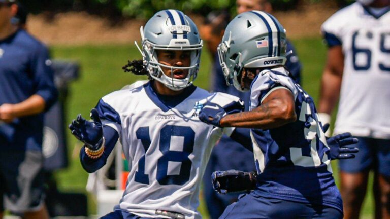 Two football players in Dallas Cowboys uniforms and helmets, including Jalen Tolbert, practice on the field with teammates in the background.