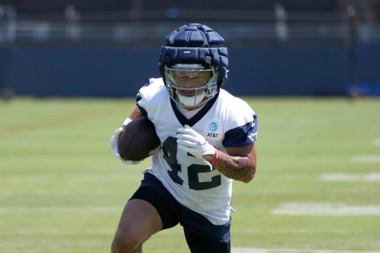 Cowboys' Deuce Vaughn, in training gear, runs with the football during a practice session on the grass field.