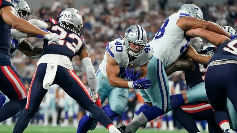 Football players from opposing teams in action during a game, with one team in blue and white uniforms and the other in blue and red. Hunter Luepke of the Cowboys makes a pivotal play that has fans on their feet.