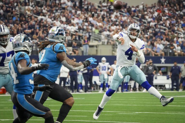 A football player in a white uniform, resembling Dak Prescott, throws a pass while three Panthers players in blue and black attempt to block the throw.