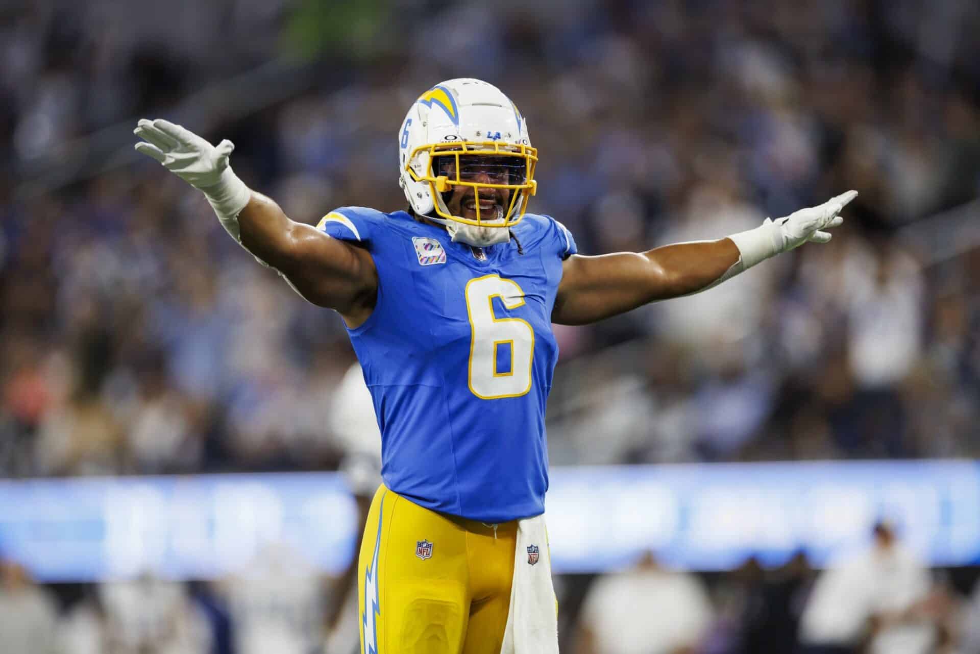 Football player in blue uniform with number 6 spreads arms wide on the field during a game. Crowd blurred in background.