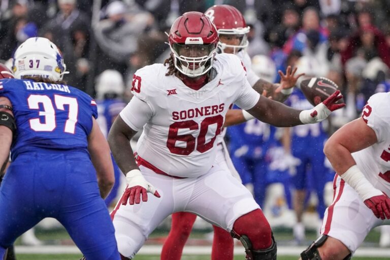 Tyler Guyton, a football player in an Oklahoma Sooners uniform, prepares to block an opponent from the opposing team wearing a blue uniform.