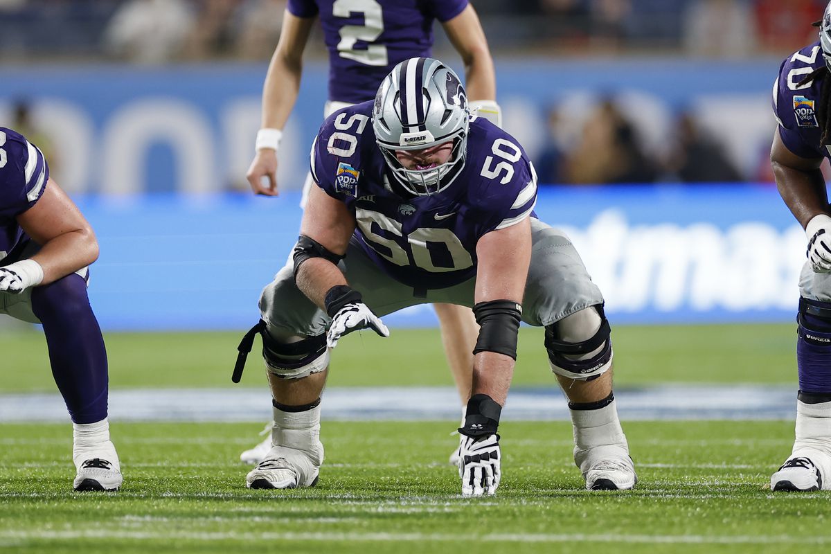 Football player in a crouched stance on the field, wearing a purple and gray uniform with the number 50. Cooper Beebe prepares to launch into action, determination evident in his eyes.