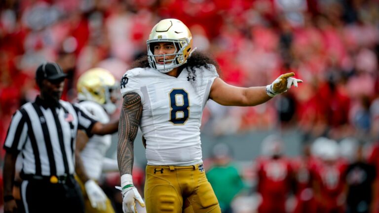 A football player in a white and yellow uniform with number 8, resembling Marist Liufau, stands on the field during a game, gesturing with his left hand.