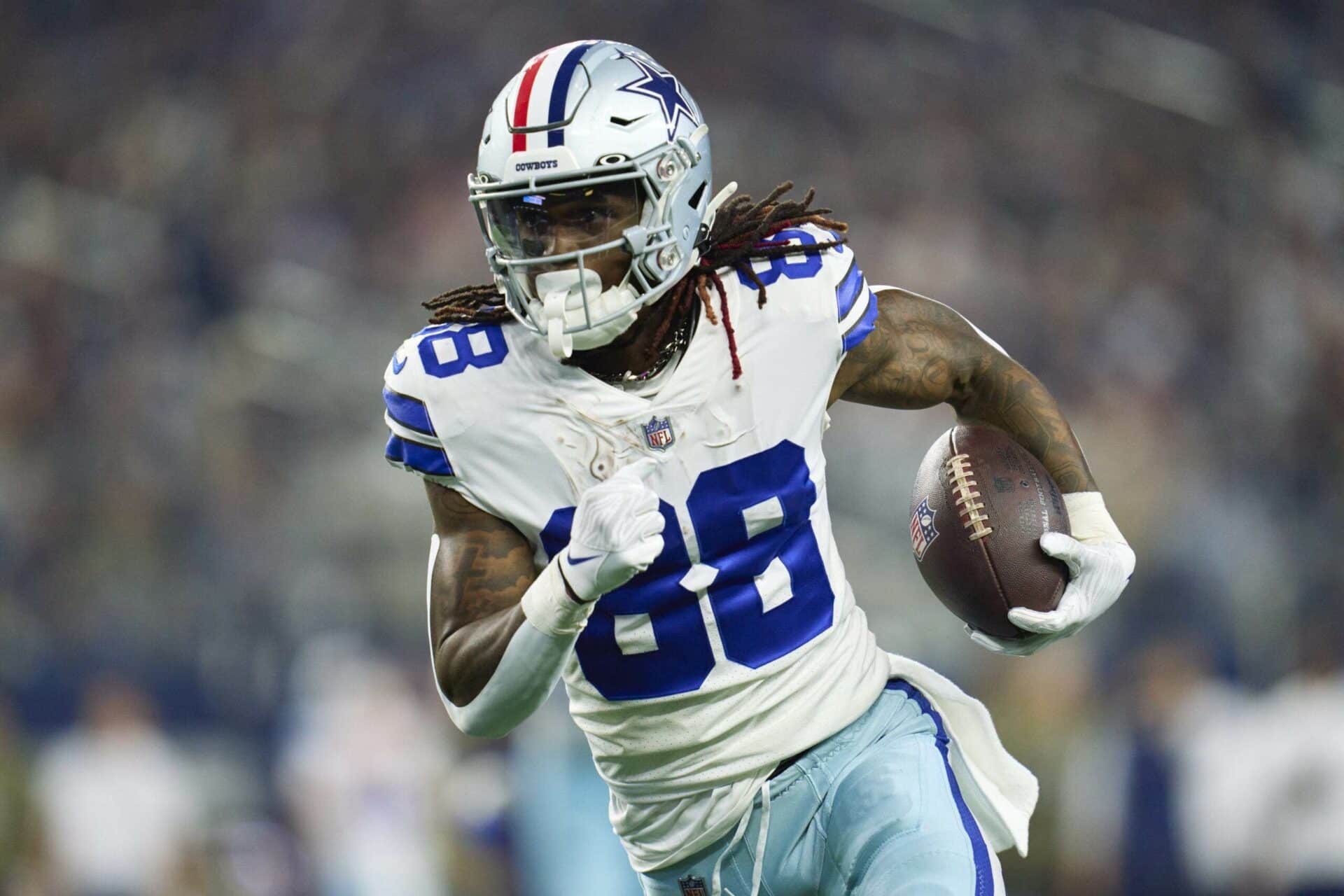 CeeDee Lamb, in a white and blue uniform, sprints determinedly across the field, clutching the football tightly during a high-stakes game.