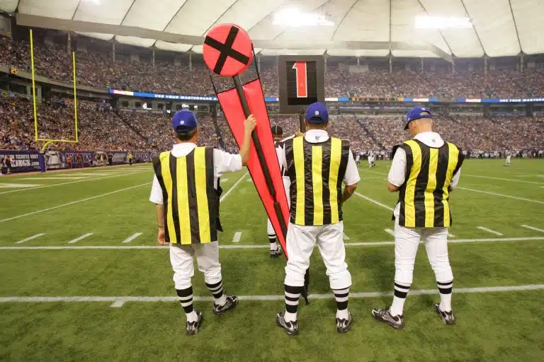 Three officials in striped uniforms hold yardage markers on the sidelines during a football game in a stadium, seamlessly integrating technology to ensure precise measurements and smooth gameplay.