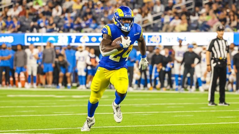 During Cowboys Training Camp, an RB in blue and yellow uniform runs with the ball during a game on a field with a crowd in the background, earning praise for his agility and performance.