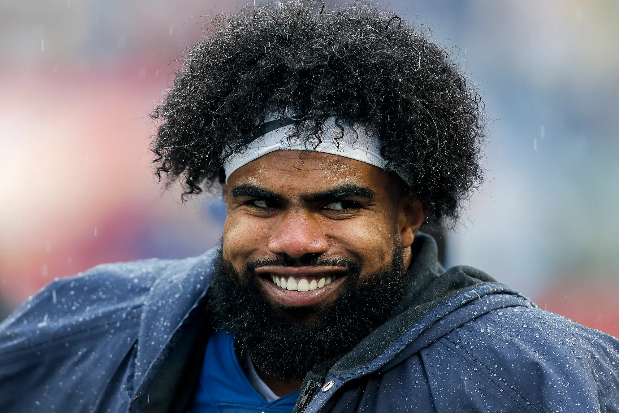 A man with curly hair and a beard, wearing a headband and a hoodie, smiles while outdoors in rainy weather.