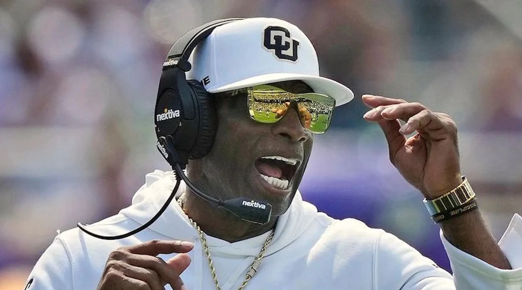 A sports coach wearing a headset and sunglasses, gesturing with one hand and speaking.