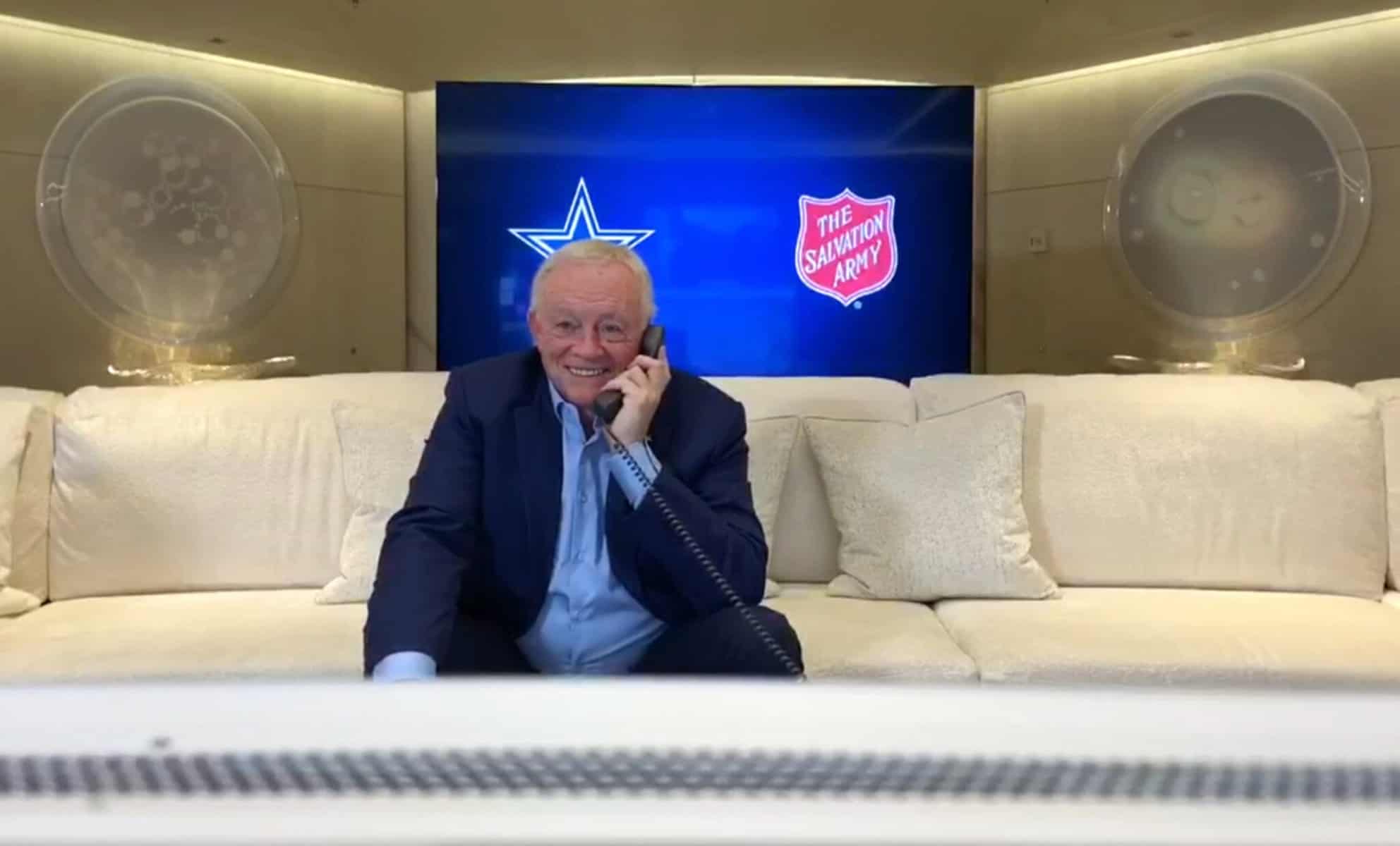 A man in a suit is sitting on a couch, talking on a phone. Behind him are logos of a football team and the Salvation Army.