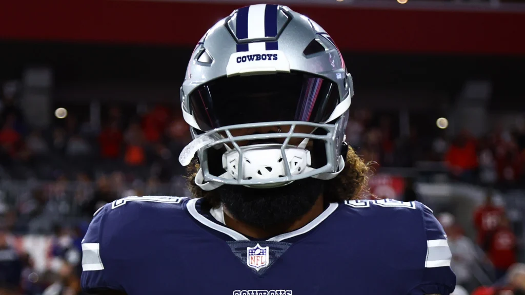 An American football player in a Dallas Cowboys uniform, wearing a helmet with a dark visor, stands on the field.