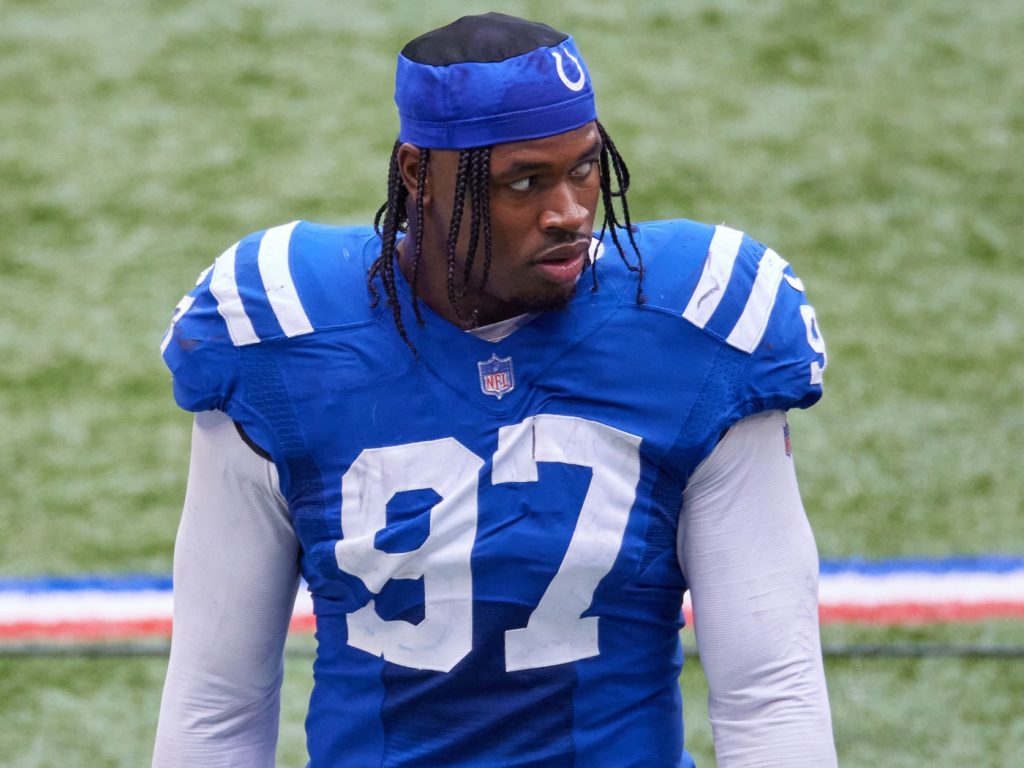 Football player wearing an Indianapolis Colts uniform with the number 97 stands on the field, looking to the side. Meanwhile, news breaks that the Cowboys sign 2 defensive ends, stirring excitement in the football community.