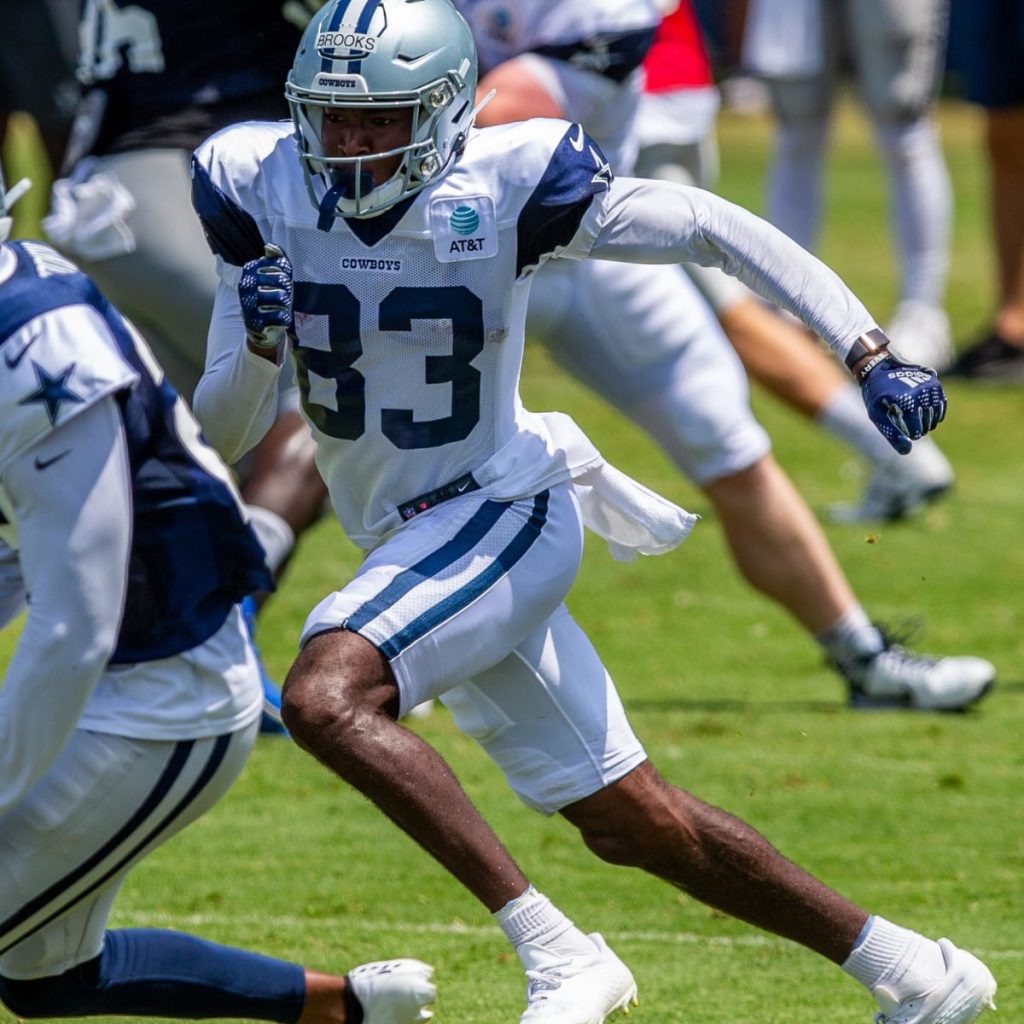 Jalen Brooks, donning a white jersey and helmet, runs during a Cowboys practice session on a grass field.