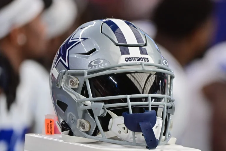 A Dallas Cowboys football helmet with a faceguard and visor, resting on a stand.