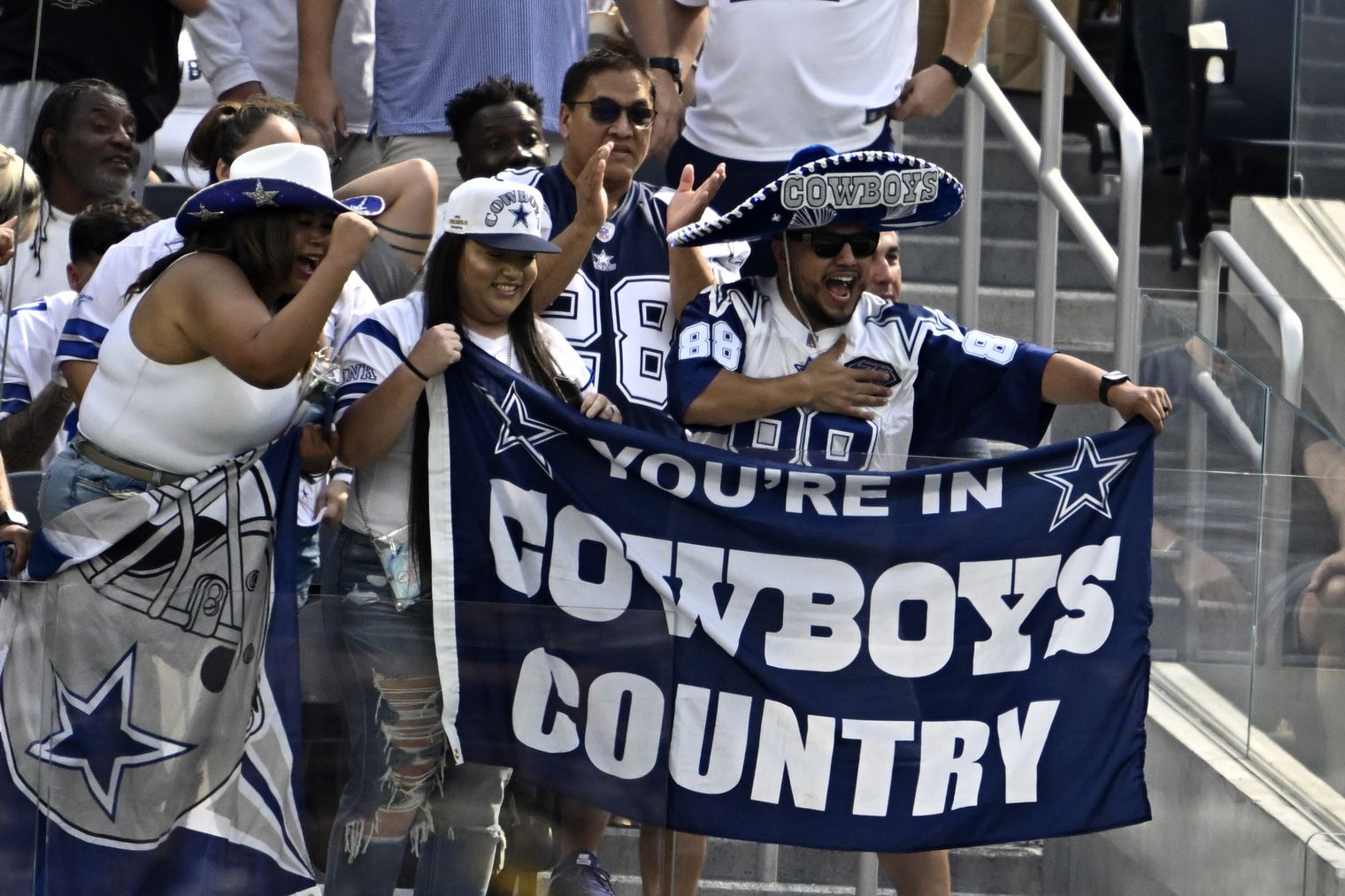 Fans wearing Cowboys gear hold a "You're in Cowboys Country" banner at a football game, cheering enthusiastically.